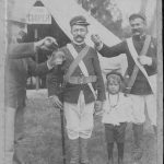 “COME HOME WITH ME FATHER” Early day photographs of Hawaii’s military clowning in uniform are practically non-existent. In this rare picture, Corporal Paul Smith (Right) of the national guard is seen playfully shaking his clenched right fist at a thirsty private. The non-coms gun barrel is reversed and hat is askew he glances sideways with anticipation at a civilian pouring him a glass of beer from a quart bottle. His trusting young son grasps the free left hand of his father, a tableau reminiscent of the old pot-boilers: the wayward, drunken parent and tearful waif pleading, “Come home with me, father!” It is 1894 and Hawaii’s first regiment is in training at camp cooper, having learned during the late 1880’s, with the Honolulu Rifles, how to “make King David Kalakaua listen to reason.”