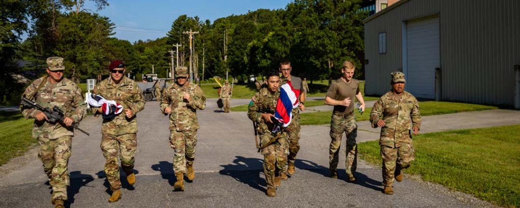 U.S. Army Spc. Andruw Nakamura, center, an infantryman assigned to Charlie Troop, 1st Squadron, 299th Cavalry Regiment, 29th Infantry Brigade Combat Team, Hawai‘i Army National Guard, finishes the 12-mile ruck final event holding the Hawai‘ian flag with supporters from Region VII during the 2024 Army National Guard Best Warrior Competition at Camp Johnson, Colchester, Vermont, Aug. 8, 2024. The Army National Guard Best Warrior Competition is a five-day event composed of 14 competitors representing the regional winners; the competition tests Soldiers’ physical and mental prowess with a series of events with the overall winners receiving either Soldier of the Year or Non-commissioned Officer of the Year recognition. (U.S. Army National Guard photo by Sgt. Lianne M. Hirano)